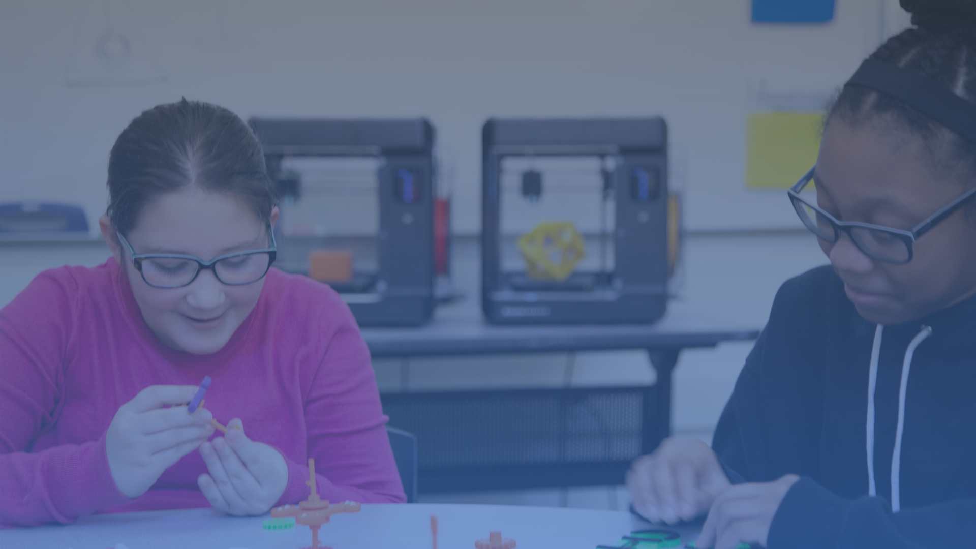 Two children with 3D printed samples and 3D printers in the background